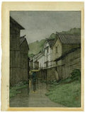 Hasui: Watercolor of an umbrella-carrying pedestrian entering a quiet village in the rain at dusk. (Sold)